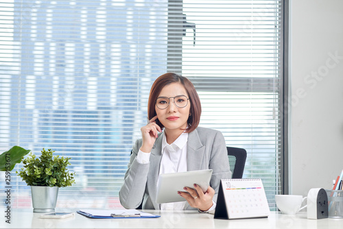 Portrait of beautiful young Asian business woman sitting and using a tablet behind the table in workplace © makistock