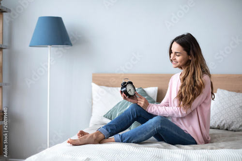 Smiling young woman lying awake in bed with alarm clock, good morning. © BestForYou