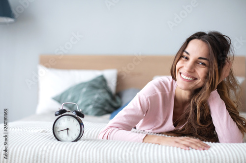 Beautiful smiling woman with alarm clock on the bed at home. © BestForYou