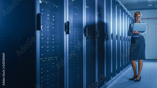 In Data Center: Female IT Technician Running Maintenance Programme on a Laptop, Controls Operational Server Rack Optimal Functioning. Modern High-Tech Telecommunications Operational Super Computer.