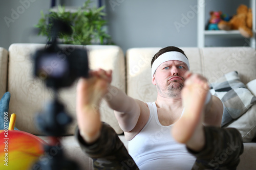 Vlogger freak shows his subscribers on camera how not to do the stretching correctly, holding his hands over his feet. Astonished face expression grimaces aganist home background concept © Hanna
