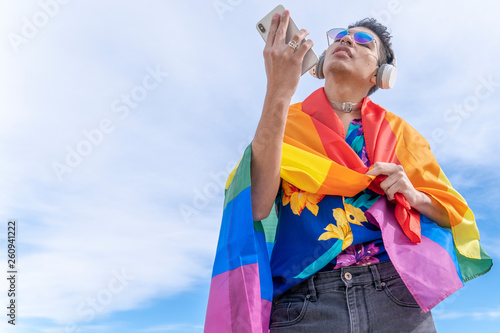 gay young man with cell mobile and flag in the wind with sky blue background © GERMAN GONZALEZ