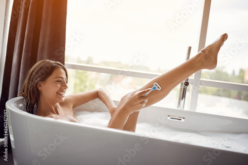 Smiling woman shaving her legs in the bath with foam. © luckybusiness
