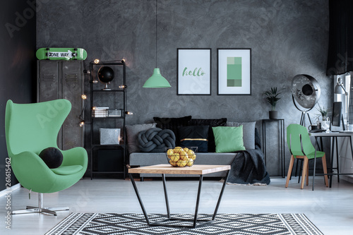 Green armchair next to wooden table in dark living room interior with posters above couch. Real photo © Photographee.eu