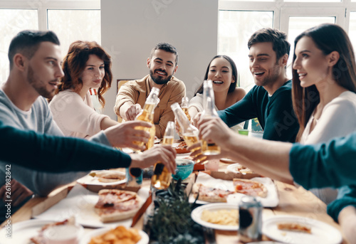 To our friendship! Group of young people in casual wear toasting each other and smiling while having a dinner party indoors © gstockstudio