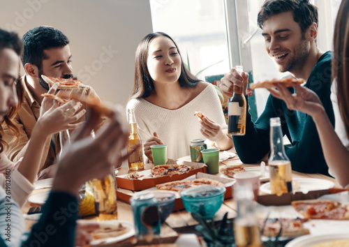 Having a great time. Group of young people in casual wear eating pizza and smiling while having a dinner party indoors © gstockstudio