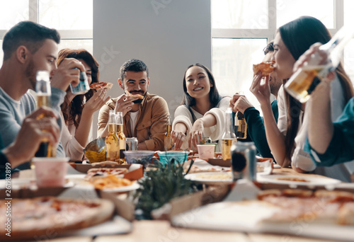 More laugh with friends. Group of young people in casual wear eating pizza and smiling while having a dinner party indoors © gstockstudio