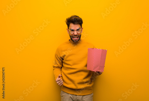 Young man holding popcorns screaming very angry and aggressive