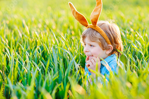 Cute little kid boy with bunny ears having fun with traditional Easter eggs hunt on warm sunny day, outdoors. Celebrating Easter holiday. Toddler finding, colorful eggs in green grass © Irina Schmidt