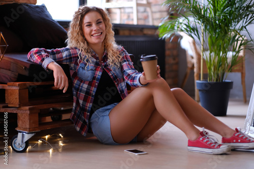 Pretty smiling woman is sitting next to the pallet sofa. She is holding cup of coffee. Woman wear hotpants and checkered shirt. Modern loft with brick wall at background. © Fxquadro