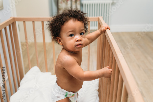 Portrait of curly haired toddler standing in a crib, looking at camera © Artem Varnitsin